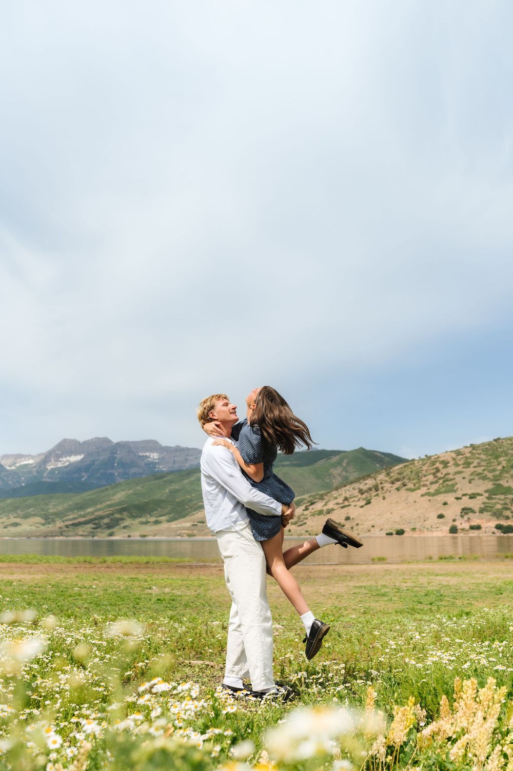 Romantic couple sharing a joyful moment as man lifts woman in a sunny wildflower field with mountain lake backdrop.