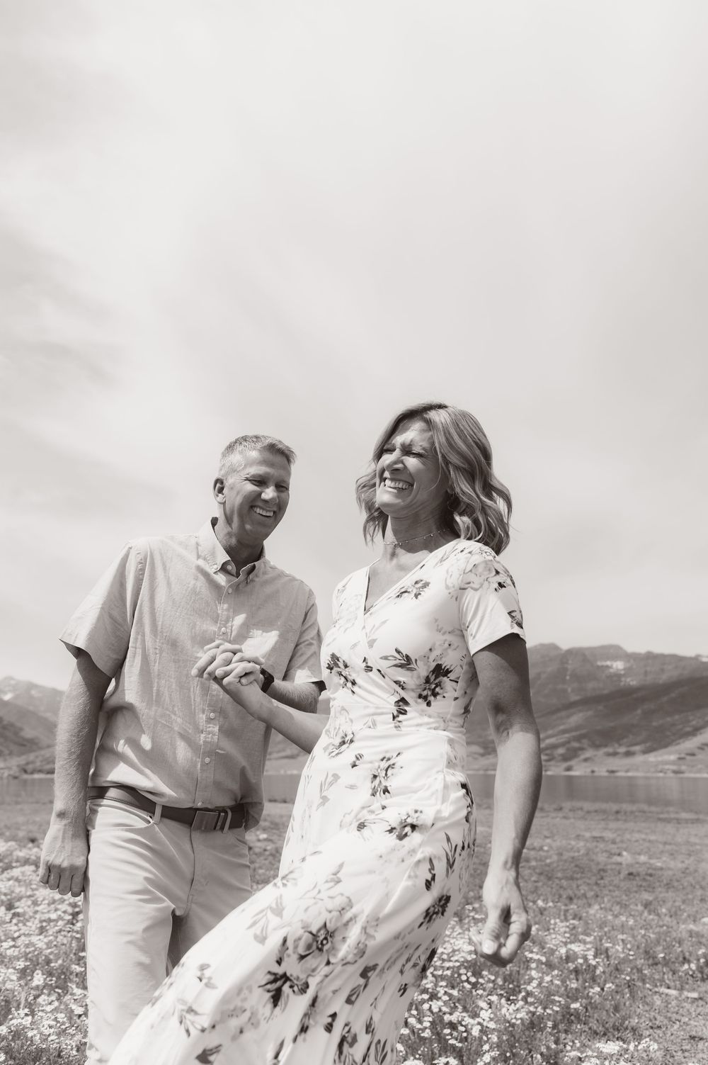 Black and white photo of a happy couple holding hands and walking through a wildflower meadow with mountains behind.