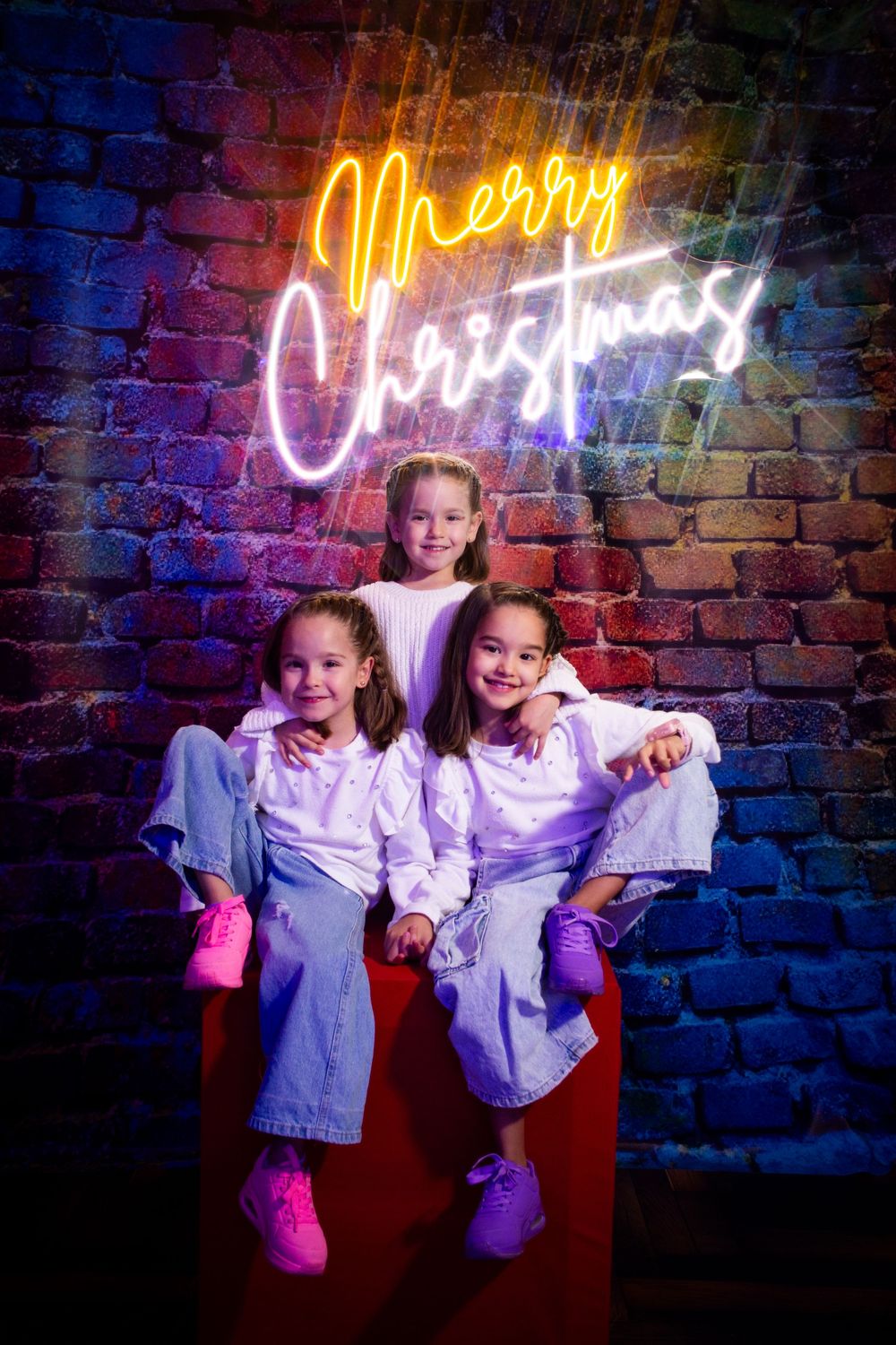 Illuminated neon Christmas text on brick wall creates festive backdrop for holiday photo shoot.
