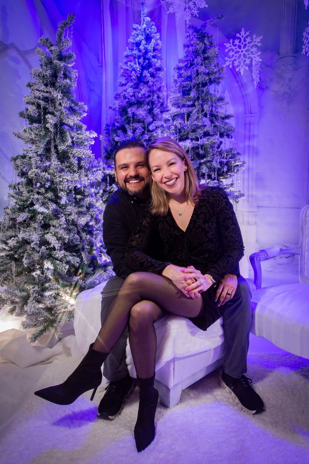 A couple enjoys a romantic winter photoshoot in front of snowy Christmas trees with purple lighting.