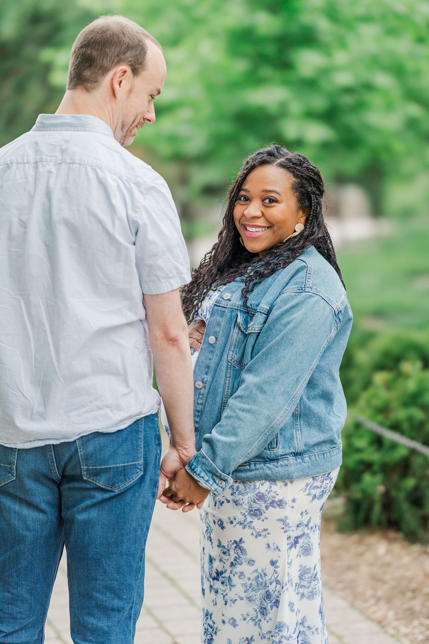 A smiling woman in a denim jacket holds hands with a man outdoors, turning back to look at the camera.