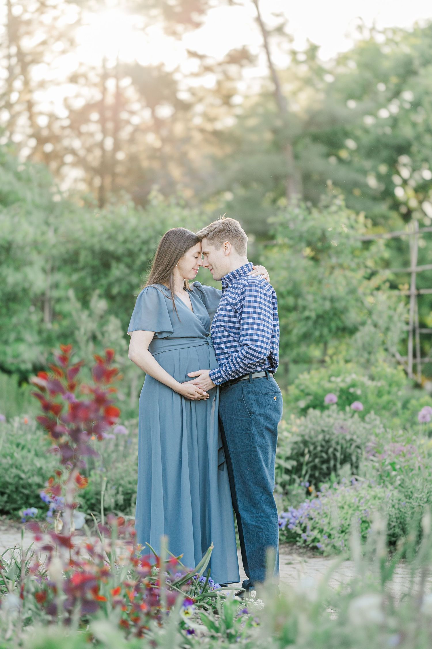 Pregnant couple embracing in a lush garden, woman in blue maternity dress, man in plaid shirt, golden sunlight.