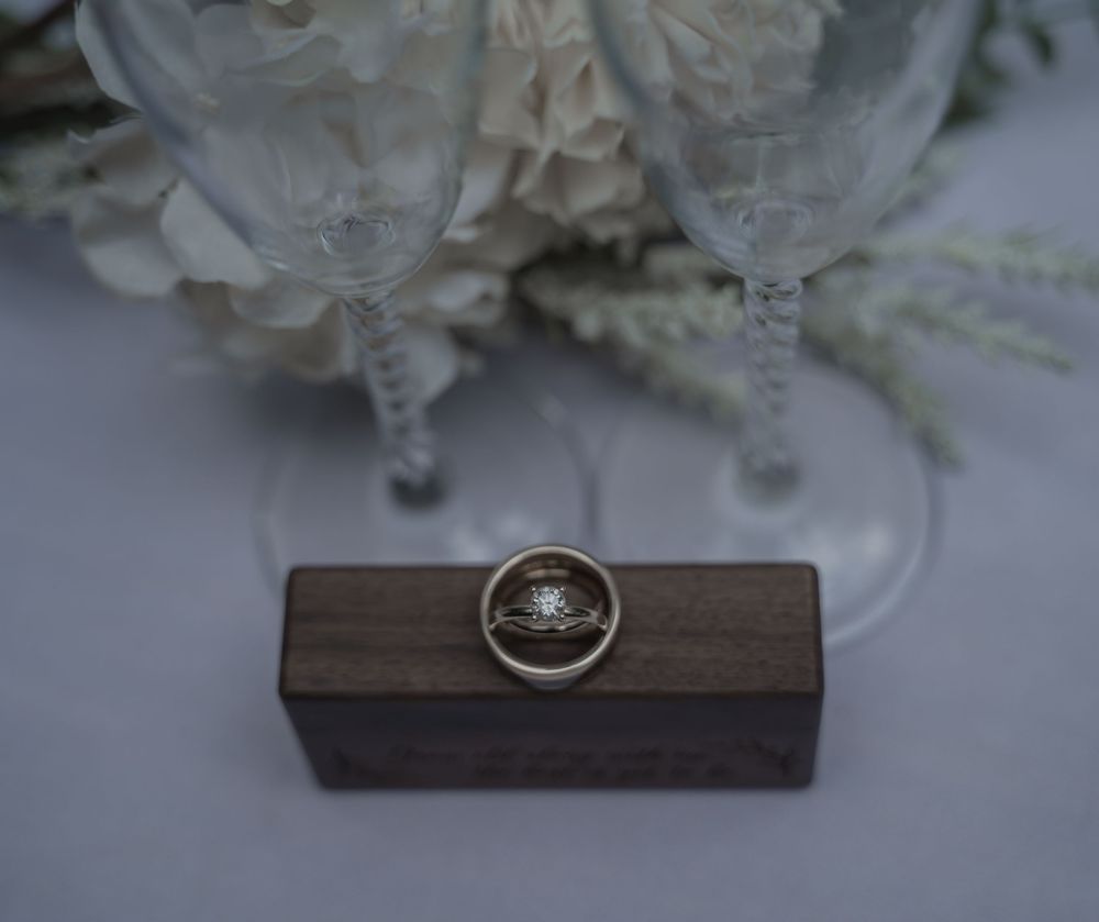 Gold diamond engagement ring displayed on a wooden ring box with crystal champagne flutes and white flowers in background.