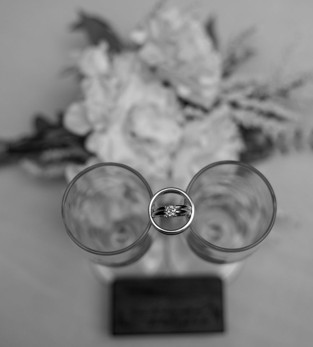 Black and white photo of wedding rings balanced between two glasses with a floral bouquet in the background.