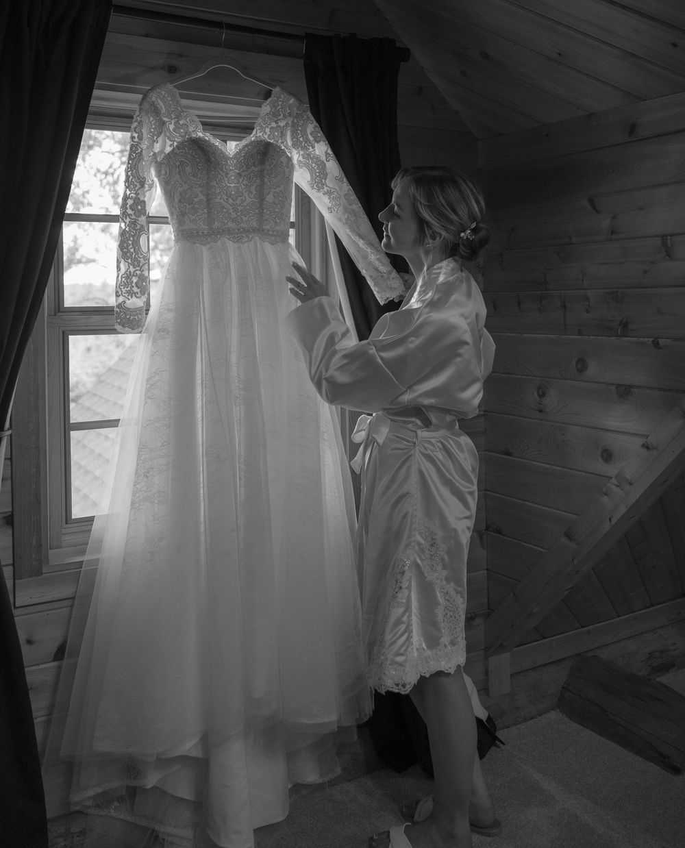 Bride in satin robe admiring her lace wedding gown hanging by a window in a rustic wooden cabin.