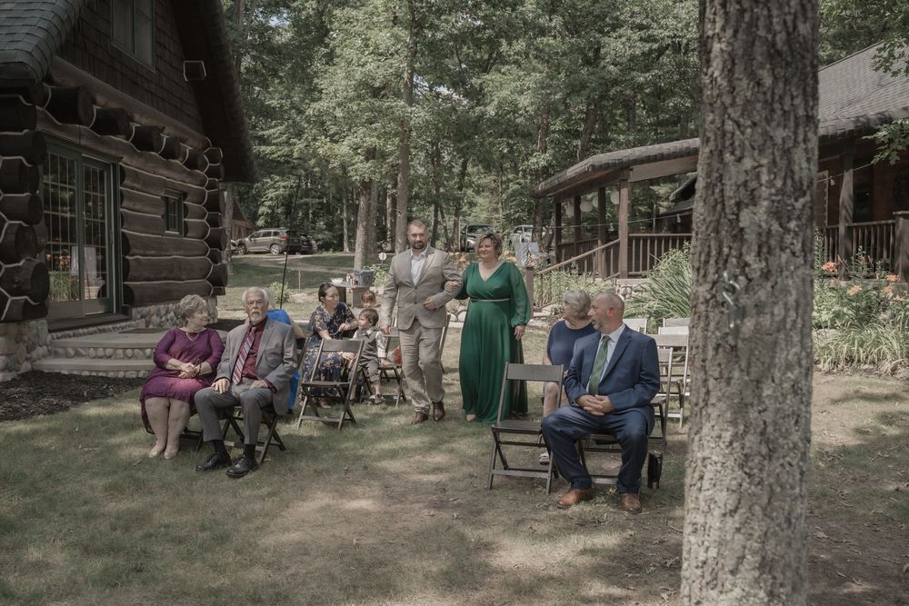 Wedding guests gathered outdoors near a rustic log cabin surrounded by tall trees for an intimate celebration.