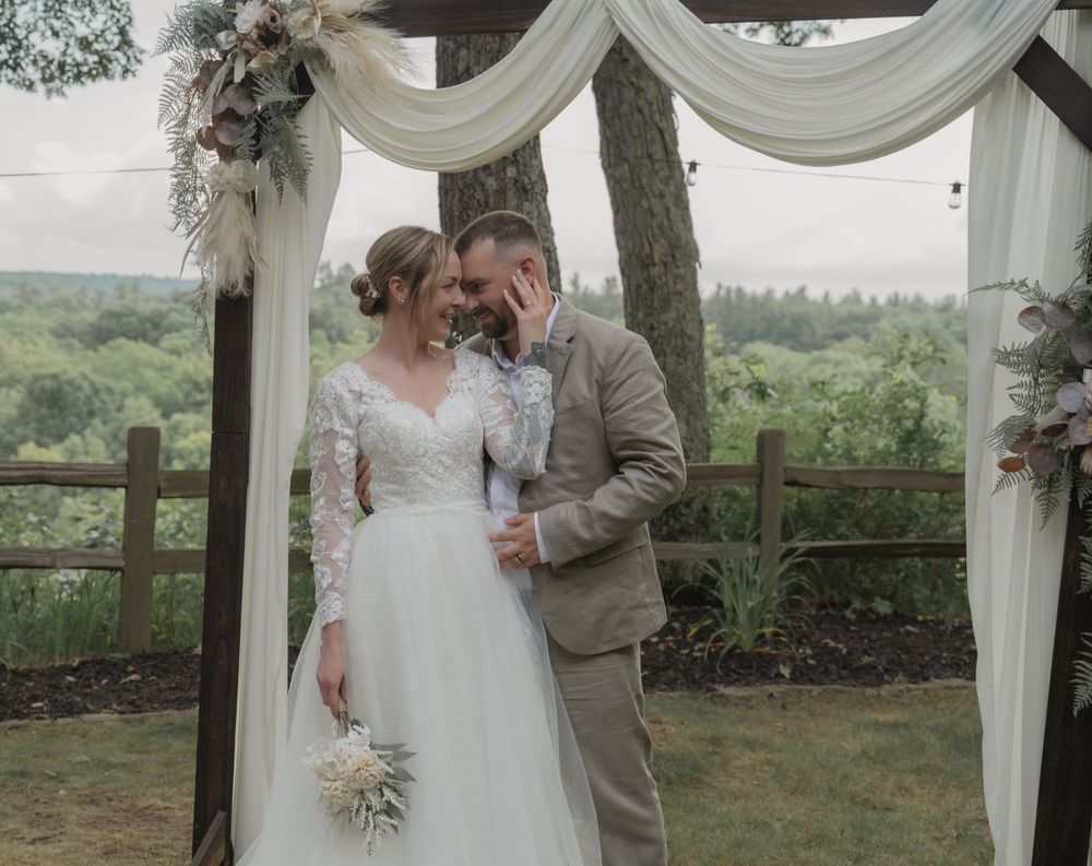 A bride and groom share a romantic moment under a floral arch at an outdoor wedding ceremony.