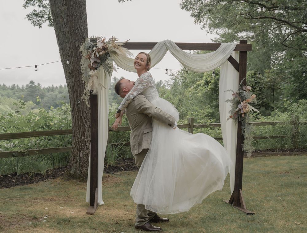 Groom lifts bride joyfully under a decorated wooden arch at an outdoor garden wedding.