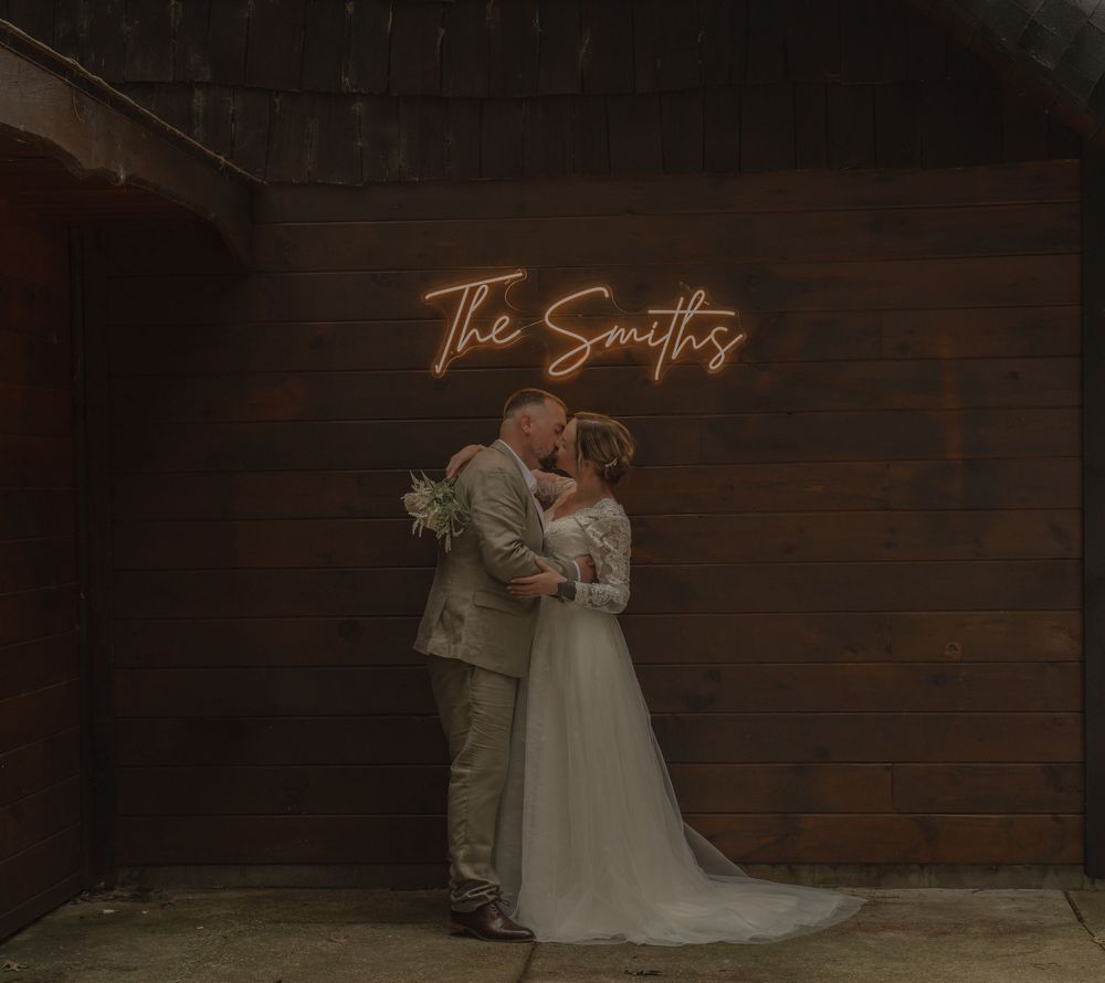 Bride and groom share a romantic kiss beneath a glowing neon sign reading The Smiths on a rustic wooden wall.