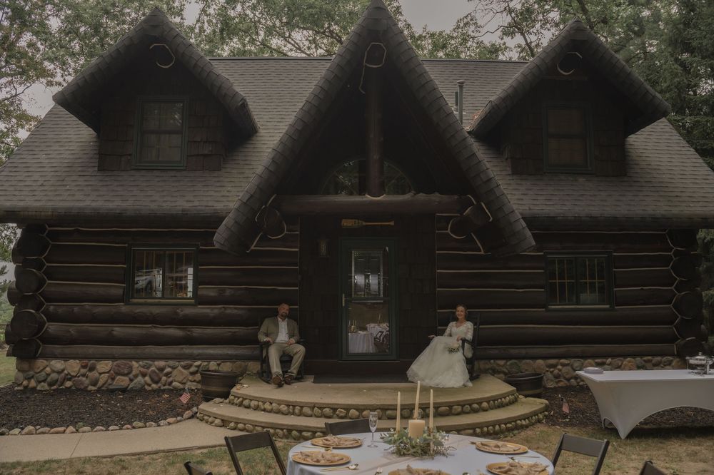 Bride and groom sitting on steps of rustic log cabin with outdoor reception table set in foreground.