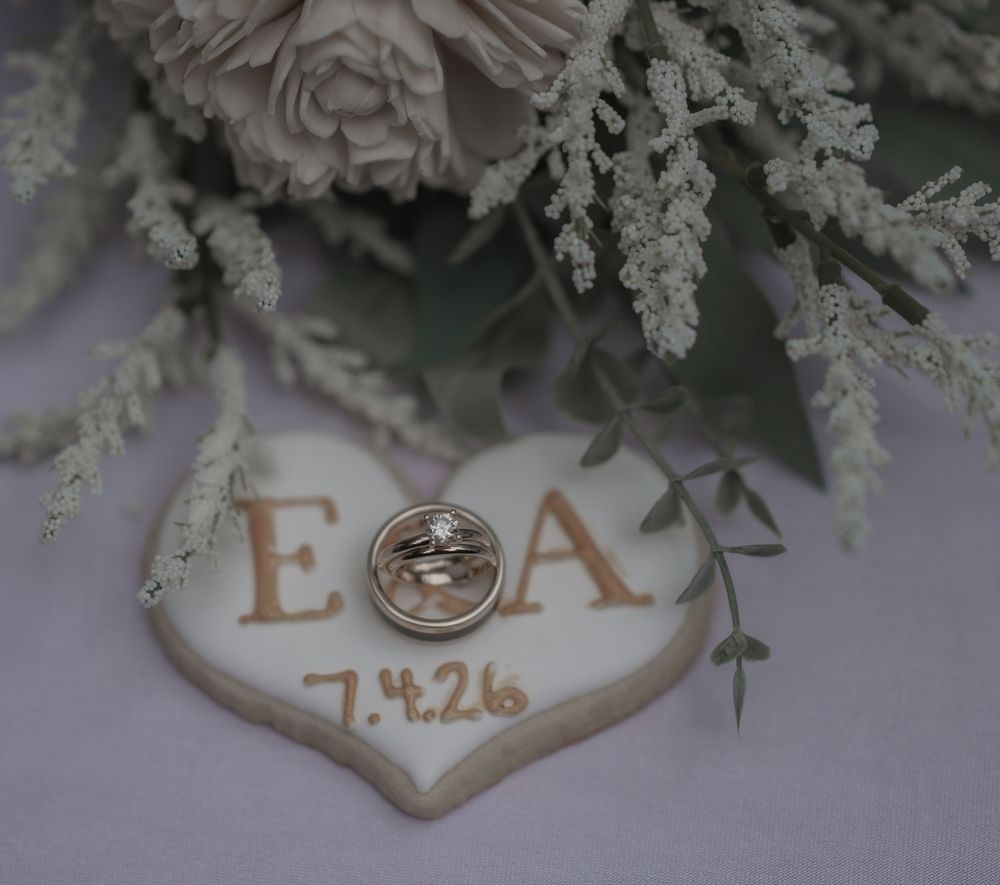 Heart-shaped wedding cookie with initials E and A and date 7.4.26, displayed with rings and floral bouquet.