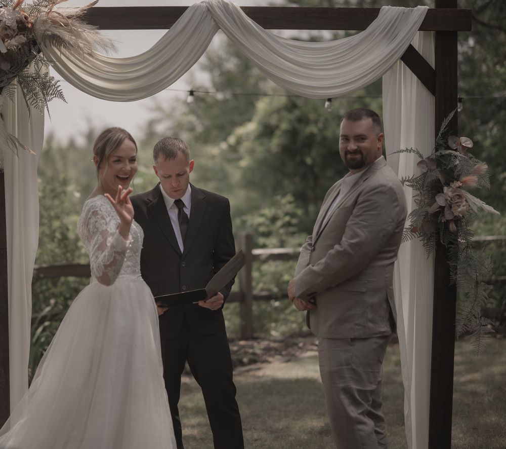 Bride and groom exchange vows under a draped wooden arch with an officiant at an outdoor wedding ceremony.