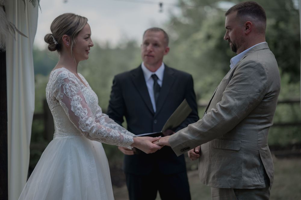 Bride and groom holding hands during outdoor wedding ceremony with officiant reading from book in background.