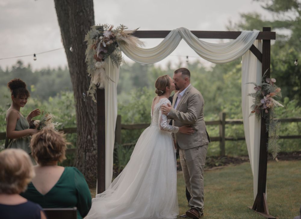 Bride and groom share first kiss under draped wooden wedding arch outdoors with lush green hills behind.
