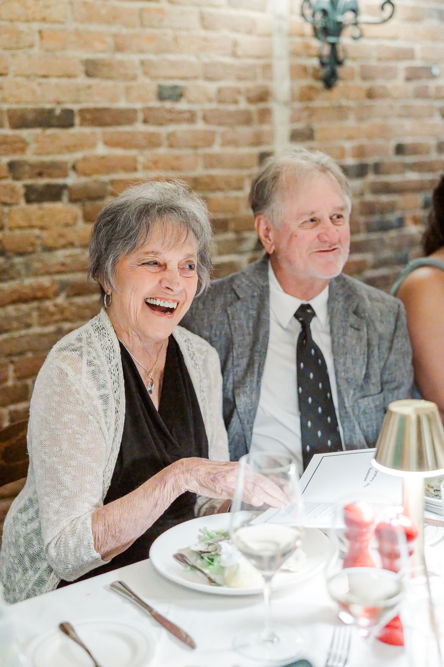 Elderly couple laughing and smiling at a formal dinner event, seated at a white tablecloth table near a brick wall.