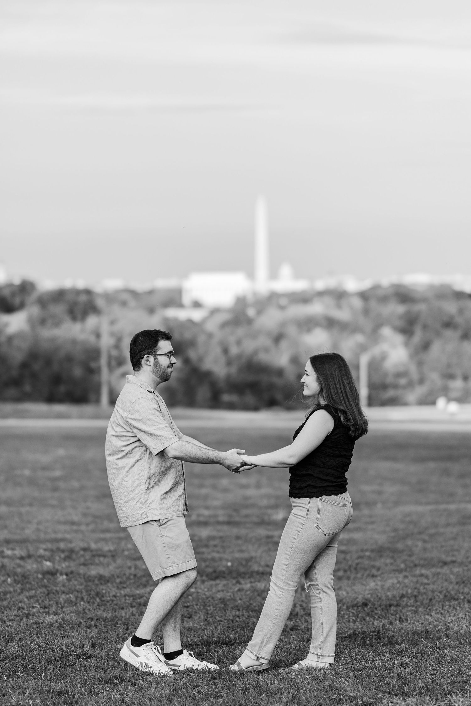 Black and white photo of a couple holding hands and facing each other on a grassy field with the Washington Monument in the background.