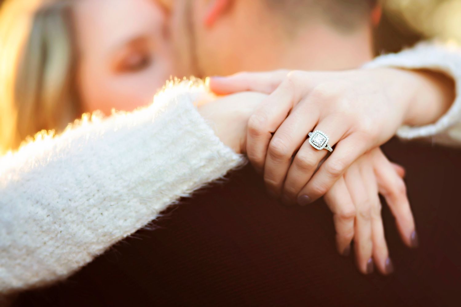 Close-up of woman's hand with diamond halo engagement ring resting on man's shoulder during romantic embrace.