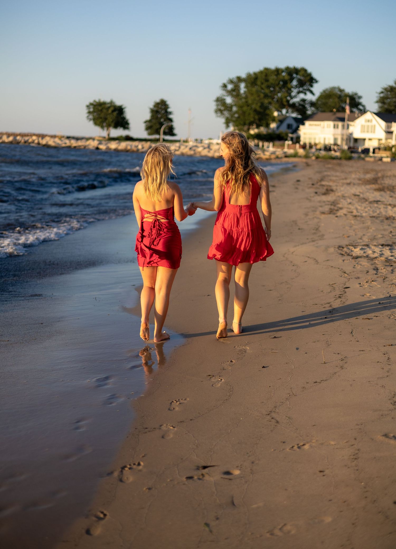 Two young women in matching red dresses walking hand in hand along a sandy beach.