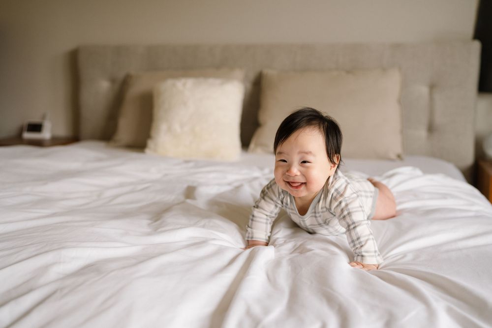 A newborn baby rests peacefully on white bedding in soft natural light.