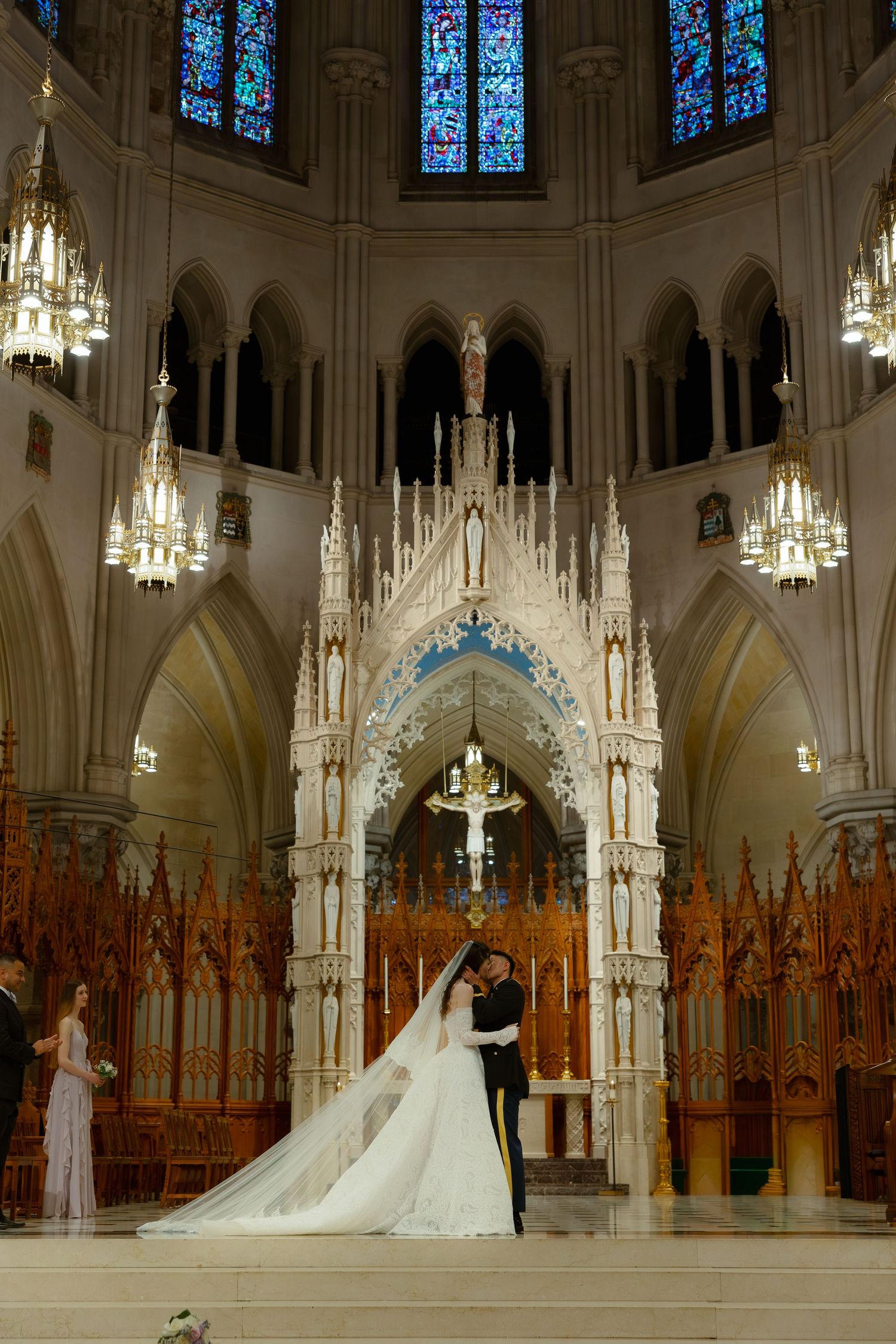 Bride and groom share first kiss at ornate Gothic cathedral altar with stunning stained glass windows and chandelier lighting.
