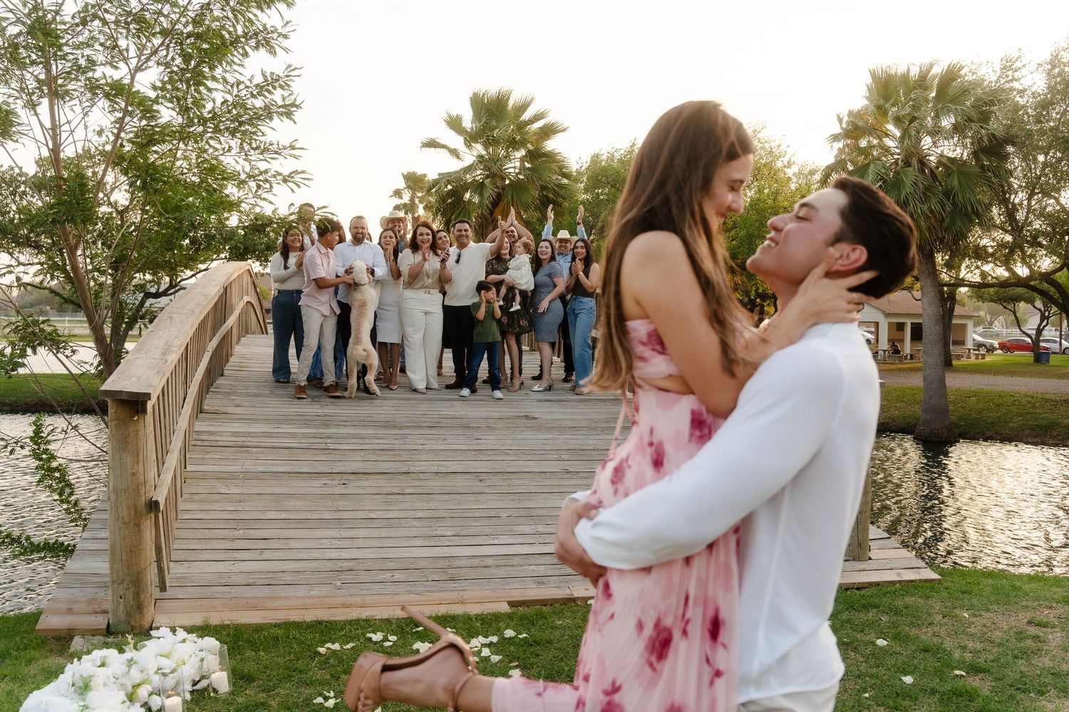 Man proposes to woman in pink dress by a wooden bridge, surrounded by cheering family and friends at sunset. McAllen Photoshoot.
