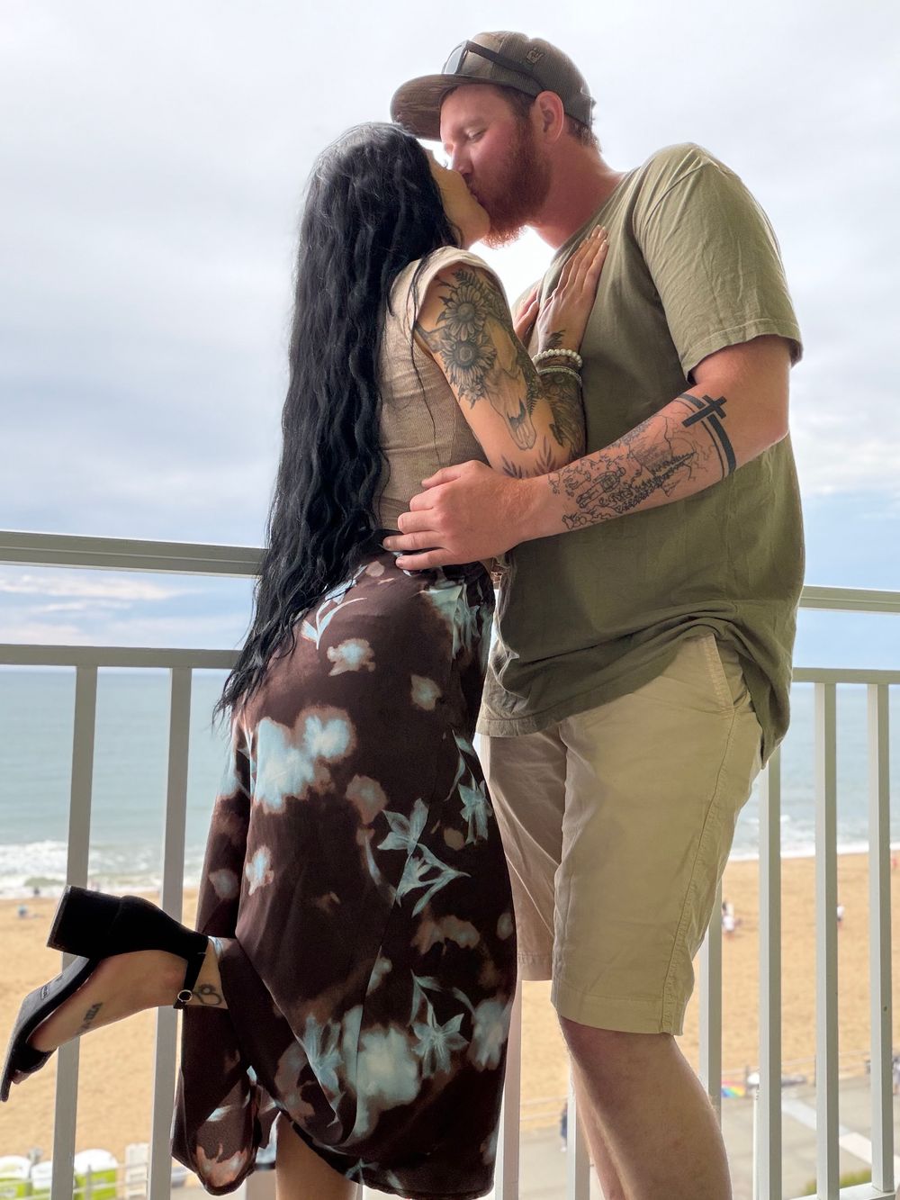 Couple sharing a romantic kiss on a seaside balcony, woman in tie-dye dress, man in green shirt with tattoos.