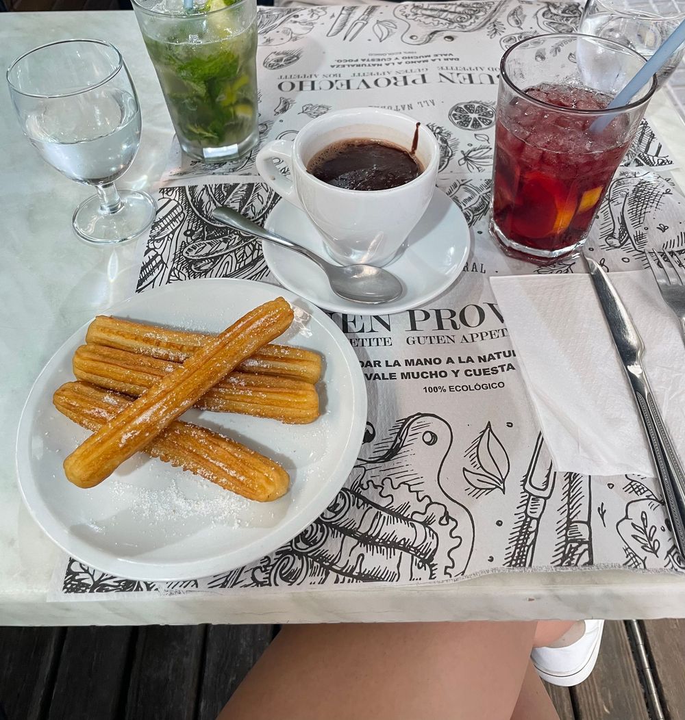 Churros with powdered sugar on a white plate, served with chocolate dipping sauce and colorful drinks on a patterned table.