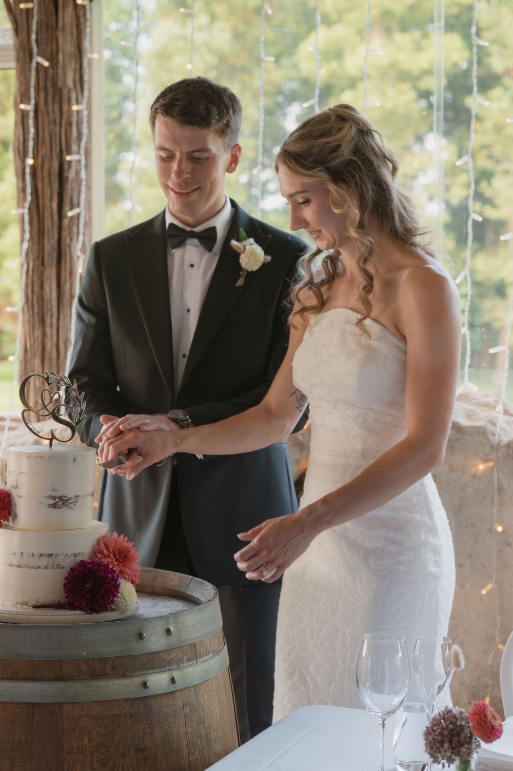 Bride and groom cutting their wedding cake on a barrel, decorated with flowers, at an elegant venue with fairy lights.