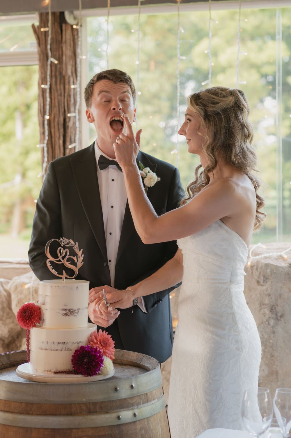Bride feeds wedding cake to groom at rustic barrel cake table, decorated with pink dahlias and fairy lights.