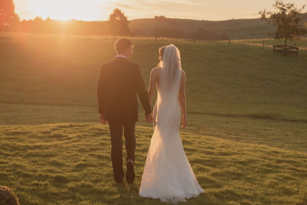 Bride and groom holding hands at sunset in a lush green field, golden hour wedding photography.