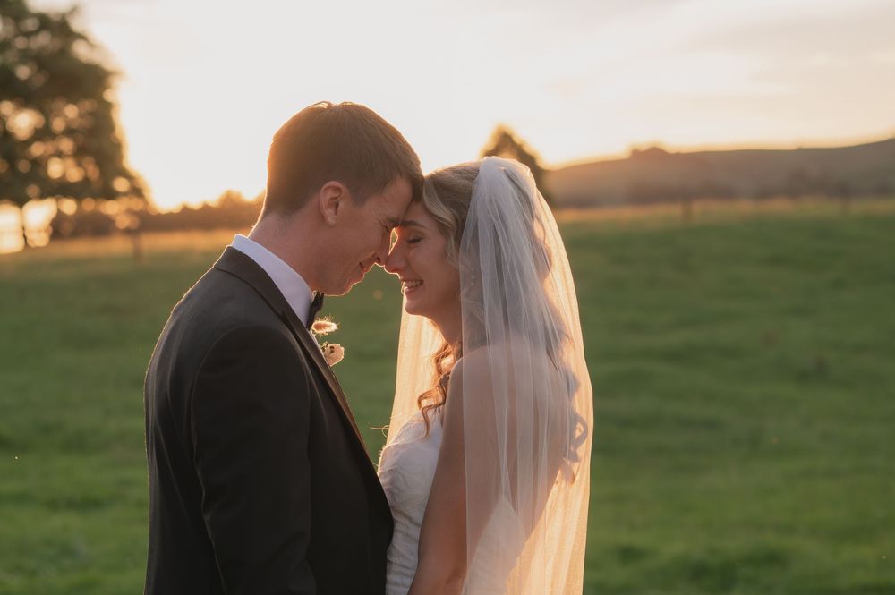 Bride and groom share a romantic forehead touch at golden hour sunset in a lush green field.