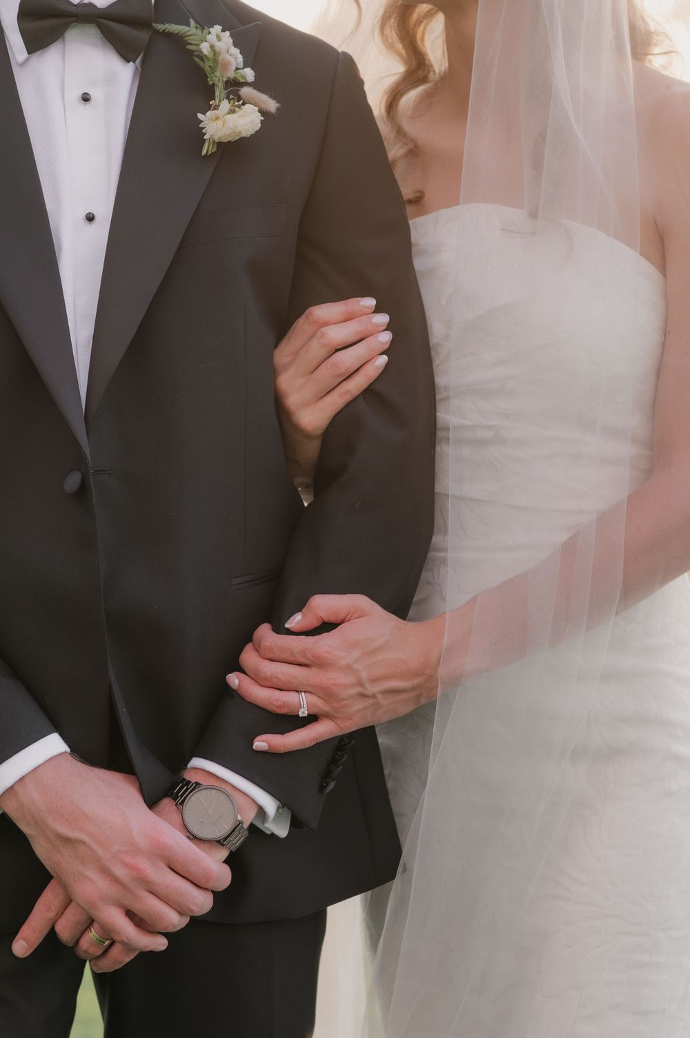 Bride and groom posing closely, she in white gown with veil, he in black tuxedo with boutonniere.