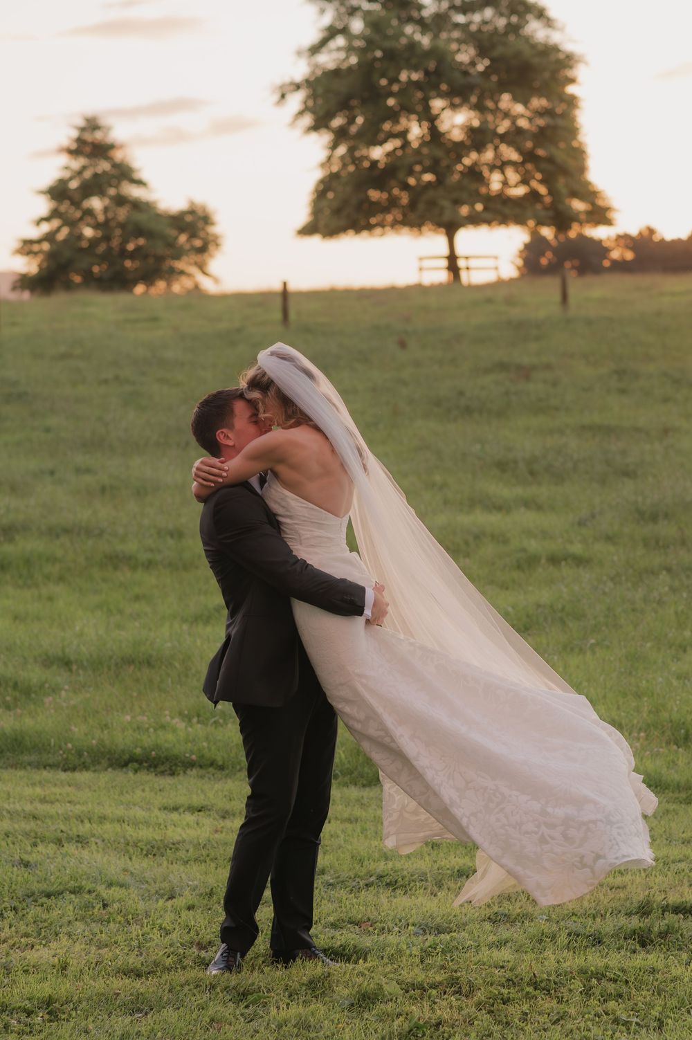 Groom lifting bride in wedding dress at sunset in a green field, sharing a romantic kiss with trees in the background.