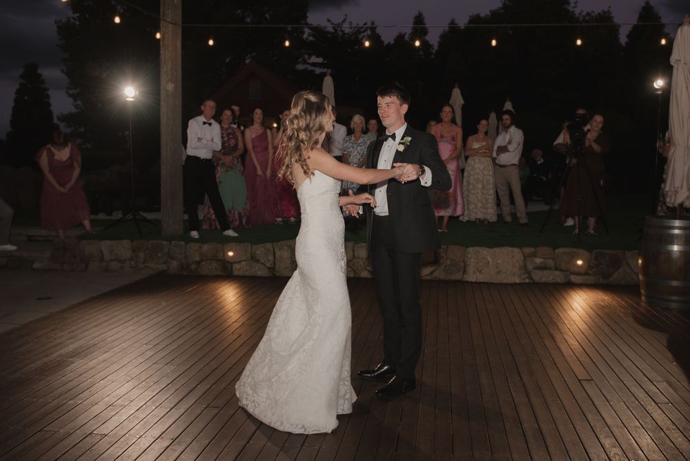 Bride and groom share their first dance on a wooden outdoor dance floor surrounded by wedding guests at night.