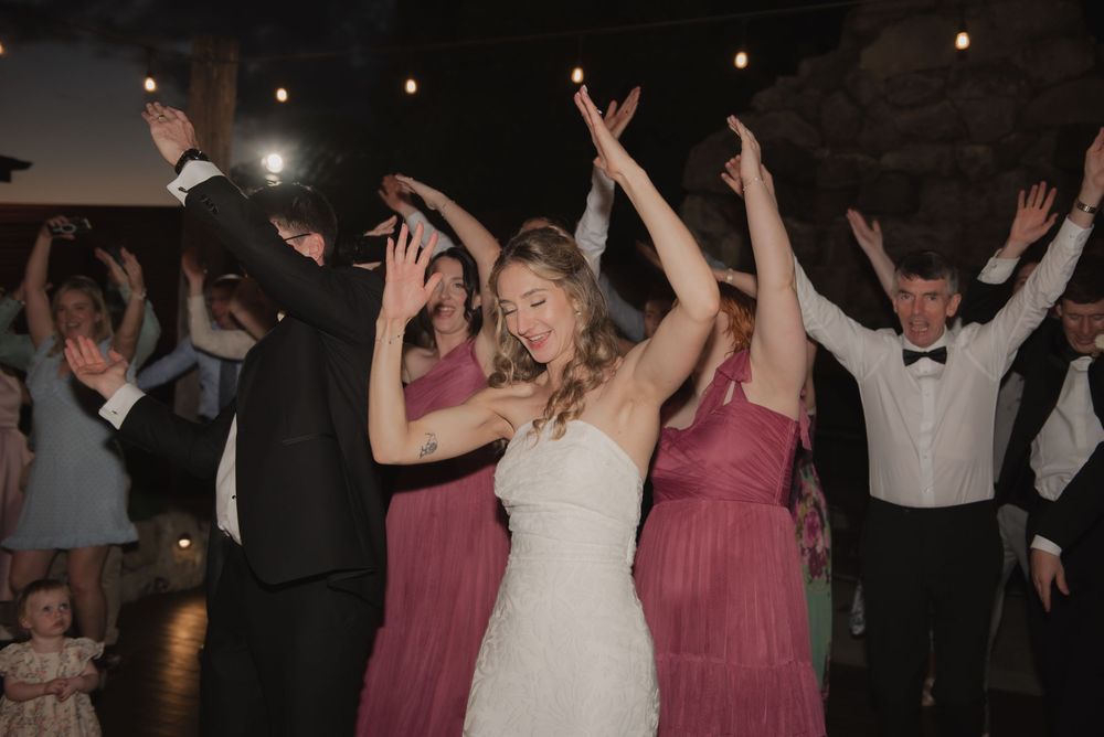 Bride in white gown dances joyfully with bridesmaids in pink dresses and guests at a wedding reception.