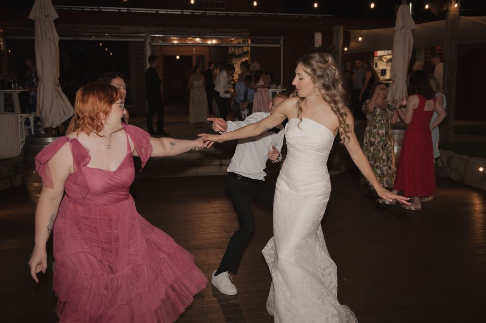 Bride in white gown dancing joyfully with guests in pink dress at lively wedding reception.