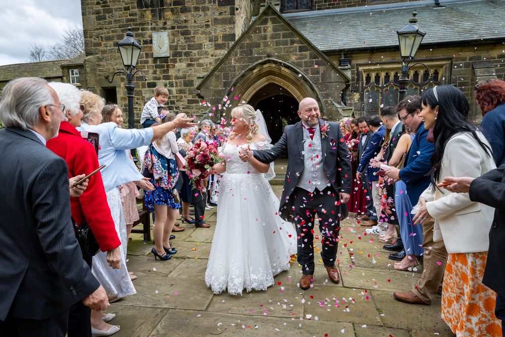 Bride and groom walk through confetti shower outside stone church surrounded by smiling wedding guests on a bright day.
