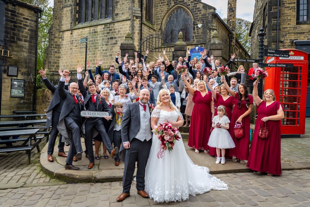Wedding party celebrating outside a stone church with bridesmaids in red, a flower girl, and guests cheering near a red phone box.