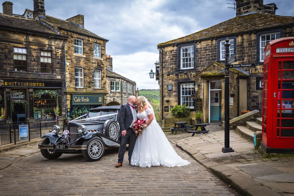 Bride and groom posing by a vintage wedding car in a charming cobblestone village square with a red phone box.