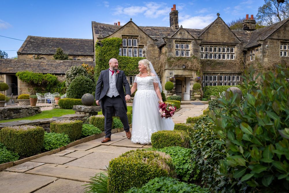 Bride and groom walking hand in hand through manicured gardens of a grand stone manor house on their wedding day.