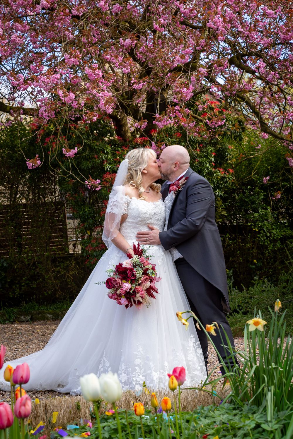 Bride and groom kissing under a pink cherry blossom tree, surrounded by colorful tulips in a garden wedding setting.