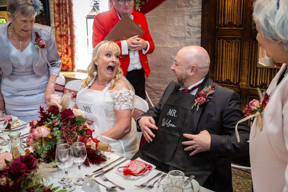 Bride and groom share a joyful moment at wedding reception table with floral arrangements and guests in background.