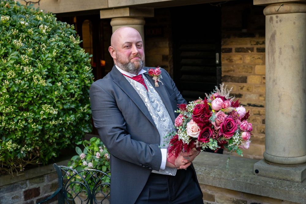 Groom in gray suit holding a vibrant bridal bouquet of red and pink roses outside a stone building.