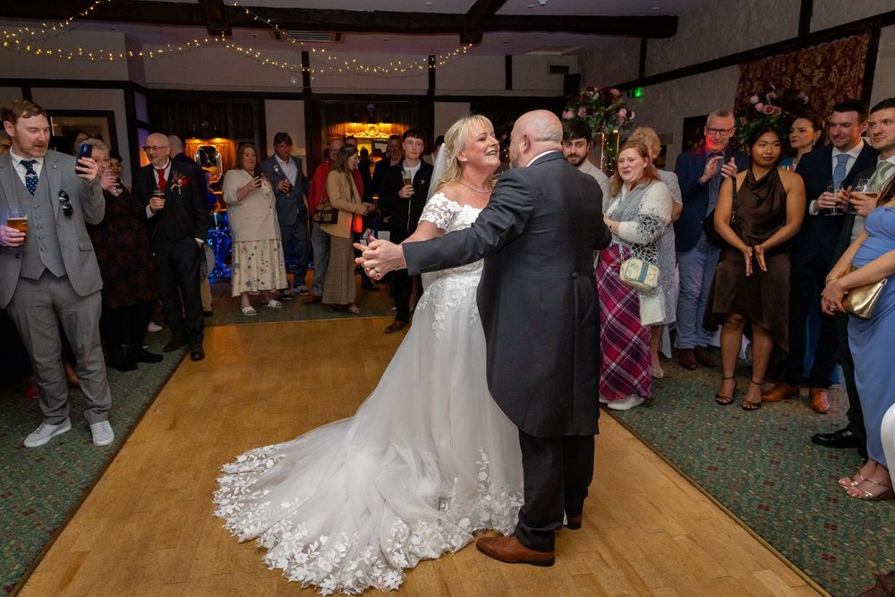 Bride and groom share their first dance at wedding reception, surrounded by guests on a wooden dance floor.