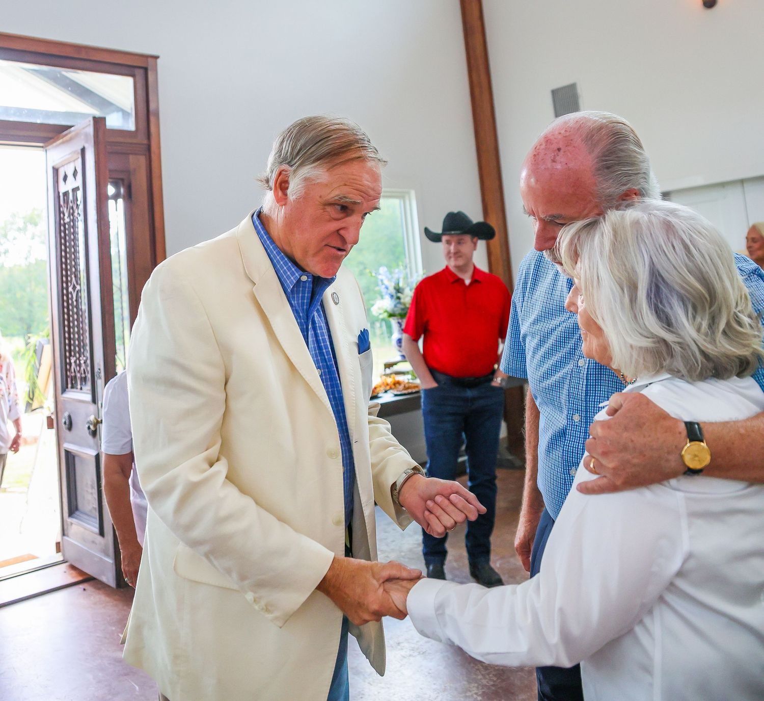 Man in white blazer shaking hands with woman at an indoor social gathering with guests in background.