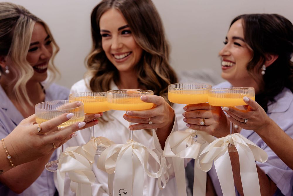 Bridesmaids in white robes toasting with orange mimosas during a bridal party getting ready celebration.
