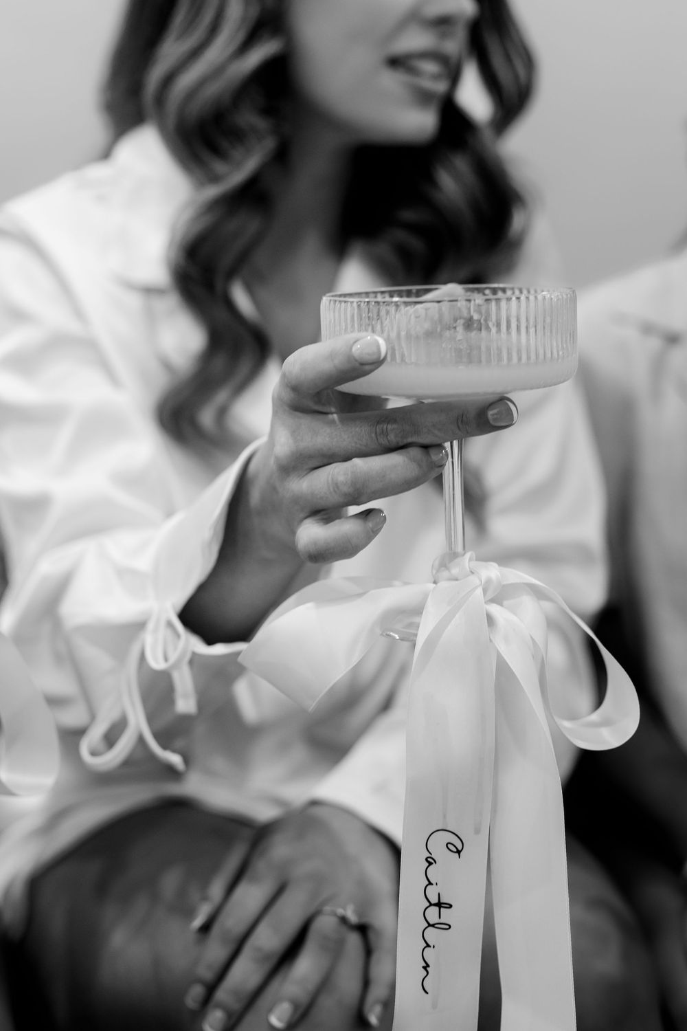 Black and white photo of a woman in a robe holding a champagne coupe glass with a ribbon labeled Caitlin.