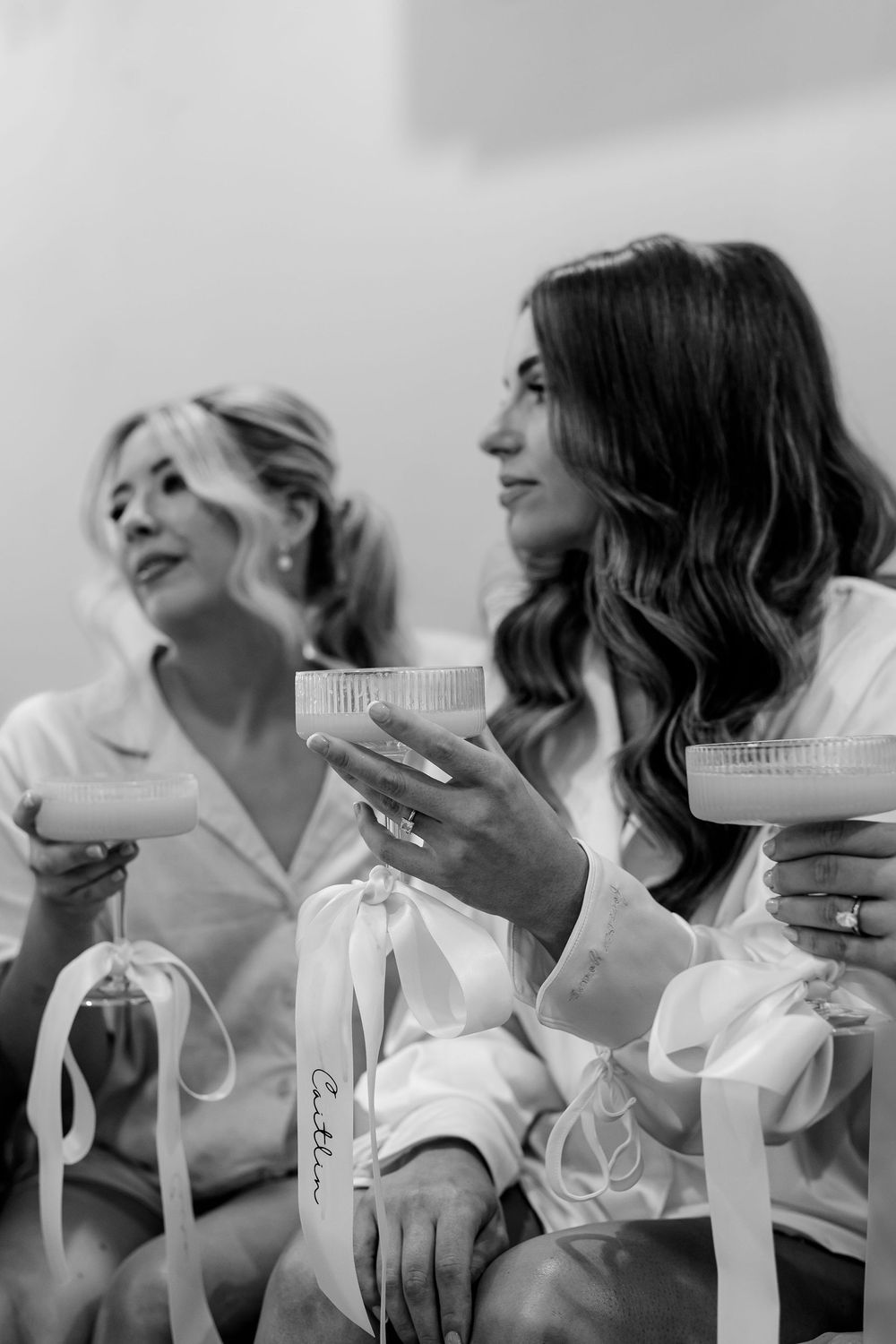 Black and white photo of two bridesmaids in satin robes holding coupe glasses during wedding morning preparations.