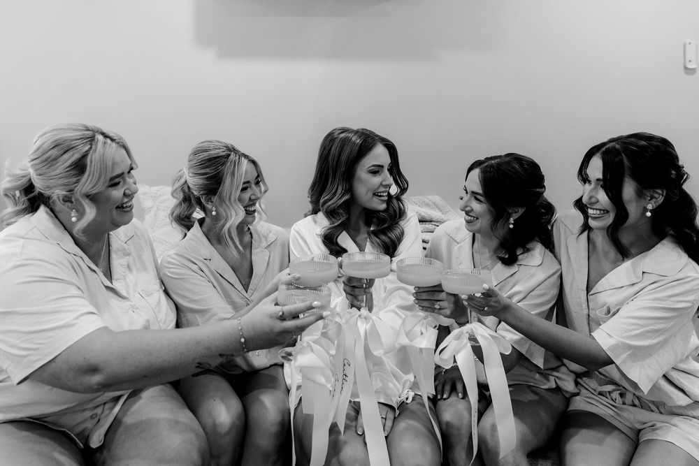 Bridesmaids in white robes toasting with champagne glasses while sitting together laughing in a bridal suite.