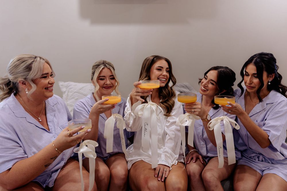 Five bridesmaids in satin robes toasting with orange cocktails while sitting together and smiling during wedding preparations.
